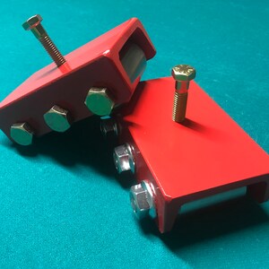 May include: Two red metal engine mounts with bolts and nuts. The mounts have a rectangular shape and are designed to support an engine.