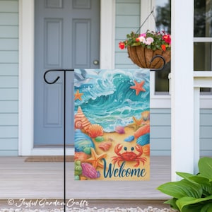 May include: A decorative garden flag featuring a beach scene with a large wave, seashells, starfish, and a red crab. The word "Welcome" is written in blue script. The flag is displayed on a black metal stand.