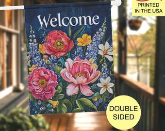 May include: A rectangular garden flag featuring a dark blue background with the word Welcome in white text. The design displays a vibrant floral arrangement of large pink and red peonies, small yellow blossoms, white lilies, and blue spikes.