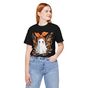 May include: Black t-shirt featuring a cute ghost wearing an orange bow and surrounded by orange flowers. The ghost has a friendly face. The shirt is paired with blue jeans. A Halloween-themed graphic tee.
