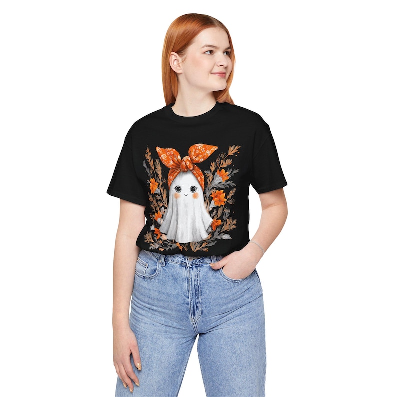 May include: Black t-shirt featuring a cute ghost wearing an orange bow and surrounded by orange flowers. The ghost has a friendly face. The shirt is paired with blue jeans. A Halloween-themed graphic tee.