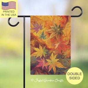 May include: A rectangular garden flag hanging on a black metal stand, featuring a vibrant autumn-themed design with multi-coloured maple leaves in shades of red, orange, yellow, and green against a swirling, abstract background of warm fall colours.
