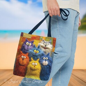 May include: A tote bag featuring a colorful illustration of six cartoon cats with expressive eyes and fluffy fur. The cats are in shades of orange, yellow, blue, and purple. The bag has a dark blue strap and the text "Joyful Garden Crafts" is visible.