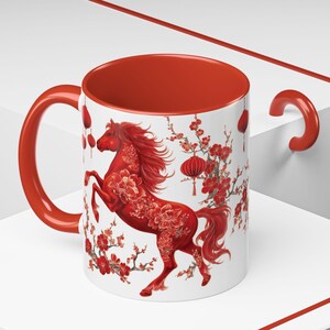 May include: A white ceramic mug with a red interior and handle. The mug features a red horse design with floral accents and hanging lanterns, set against a white background. The design is likely inspired by Asian art.