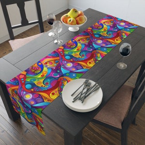 May include: A colorful table runner with a psychedelic pattern of swirls, circles, and lines in shades of blue, green, yellow, red, and purple. The runner is laid out on a dark wood table with a bowl of apples, wine glasses, and silverware.