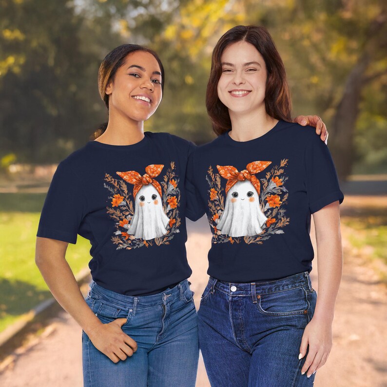 May include: Two people wearing navy blue t-shirts with a cute ghost graphic. The ghost has a white body, an orange bow, and is surrounded by orange flowers and foliage. The t-shirts are paired with blue jeans.