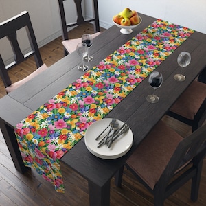 May include: A colorful floral table runner with a variety of flowers in shades of pink, yellow, red, blue, and white. The runner is laid on a dark wood table with two chairs and a set of white plates and silverware.