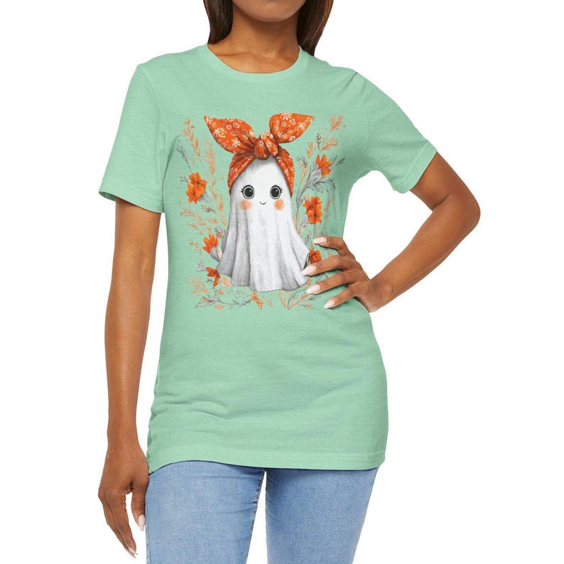 May include: Mint green t-shirt featuring a cute ghost illustration. The ghost wears a floral headband and is surrounded by orange flowers and foliage. The shirt is a short-sleeved crew neck.