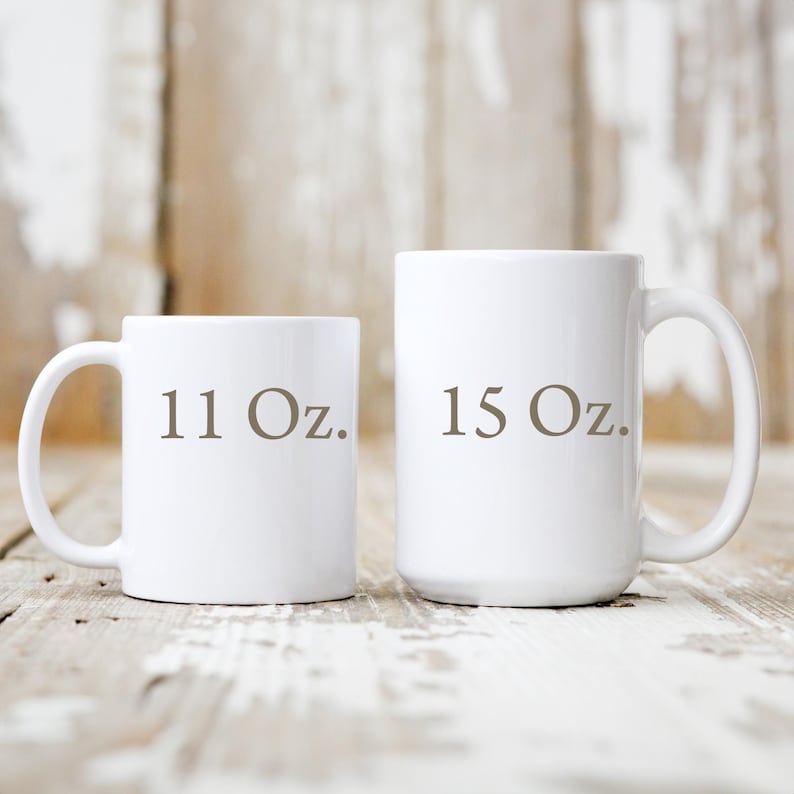 May include: Two white ceramic mugs with curved handles. The smaller mug is labeled "11 Oz." in brown text, and the larger mug is labeled "15 Oz." The mugs are set against a light-colored, weathered wooden surface.