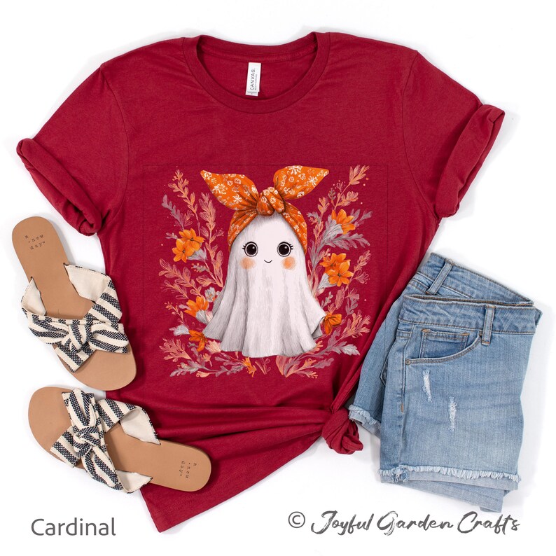 May include: A cardinal red t-shirt featuring a cute ghost illustration wearing an orange floral headband, surrounded by orange flowers and greenery. The shirt is paired with denim shorts and sandals.