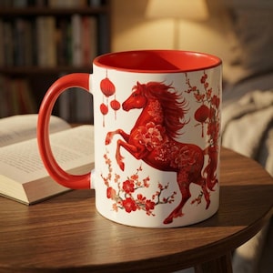 May include: A white ceramic mug with a red interior and handle. The mug features a red horse design with floral accents and hanging lanterns. The mug is on a wooden table.