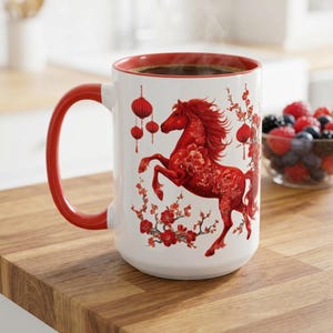 May include: White ceramic mug with a red interior and handle. The mug features a red horse design with floral accents and hanging lanterns. The mug is filled with a dark liquid, possibly coffee or tea.