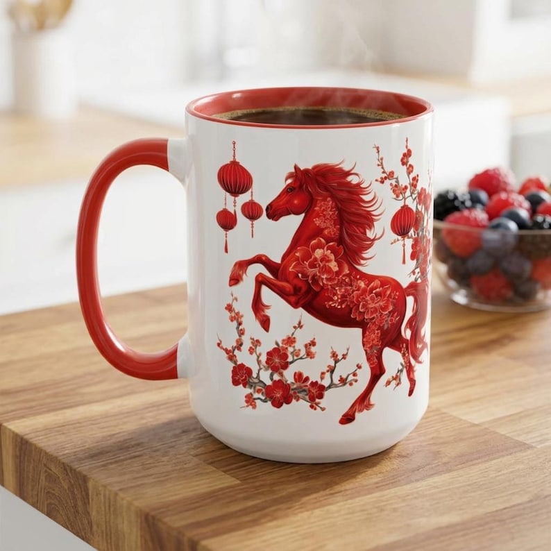 May include: White ceramic mug with a red interior and handle. The mug features a red horse design with floral accents and hanging lanterns. The mug is filled with a dark liquid, possibly coffee or tea.