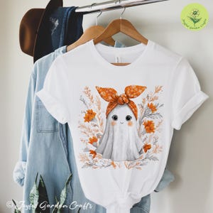 May include: White t-shirt featuring a cute ghost illustration wearing an orange bow and surrounded by orange flowers and foliage. The shirt is hanging on a wooden hanger. The text "Joyful Garden Crafts" is visible.
