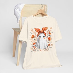 May include: Cream-colored t-shirt featuring a cute ghost illustration. The ghost wears an orange bow and is surrounded by orange flowers and foliage. A white baseball cap rests on a wooden chair in the background.