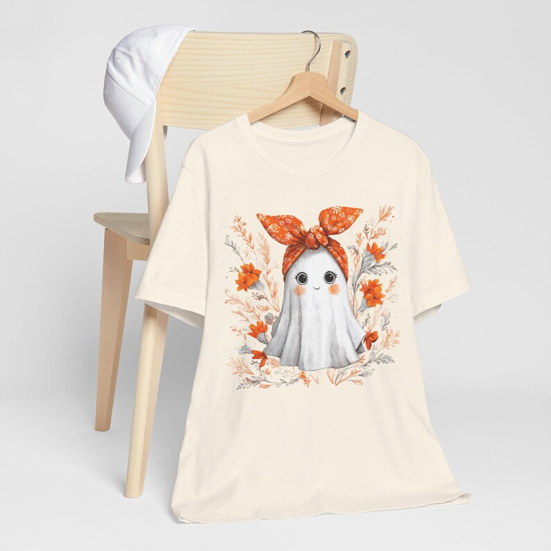 May include: Cream-colored t-shirt featuring a cute ghost illustration. The ghost wears an orange bow and is surrounded by orange flowers and foliage. A white baseball cap rests on a wooden chair in the background.