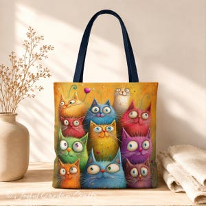 Peut inclure: Un sac fourre-tout à anses bleu foncé orné d'une illustration colorée de onze chats fantaisistes aux teintes orange, bleu, vert, rouge, violet et blanc, dotés de grands yeux expressifs et de longues moustaches sur un fond jaune chaud.