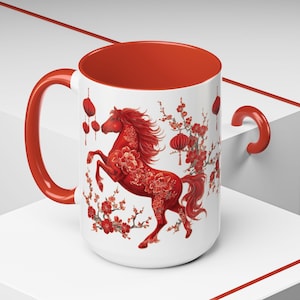 May include: A white ceramic mug with a red interior and handle. The mug features a red horse design with floral accents and hanging lanterns, set against a white background. The design is inspired by Asian art.