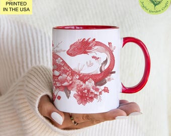 May include: White ceramic mug with a red rim and handle. The mug features a red dragon design with floral accents. The text "Joyful Garden Crafts" is printed on the bottom of the mug.