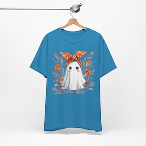May include: A turquoise blue t-shirt featuring a cartoon ghost character with a white sheet, wearing an orange bandana. The ghost is surrounded by orange flowers and green leaves. The shirt is hanging on a wooden hanger.