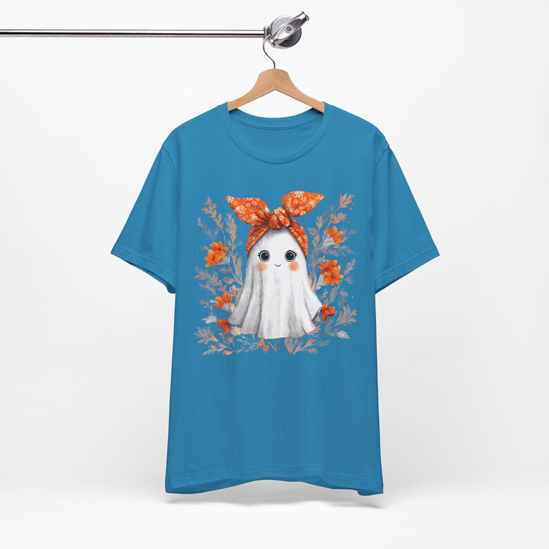 May include: A turquoise blue t-shirt featuring a cartoon ghost character with a white sheet, wearing an orange bandana. The ghost is surrounded by orange flowers and green leaves. The shirt is hanging on a wooden hanger.