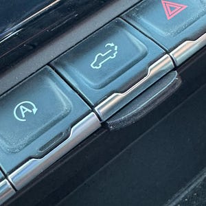 May include: A close-up of a car's dashboard with several buttons. The buttons are black with silver trim and have various symbols on them, including a car with an arrow, a triangle with an exclamation point, and a circle with an arrow.