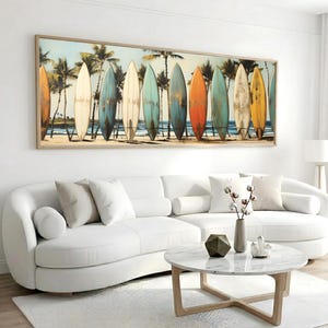 Könnte beinhalten: Ein gerahmtes Kunstwerk mit einer Reihe von Surfbrettern an einem Strand mit Palmen. Die Surfbretter sind in verschiedenen Farben gehalten, darunter Orange und Blau. Das Kunstwerk hängt über einem weißen, geschwungenen Sofa und einem runden Marmor-Couchtisch.