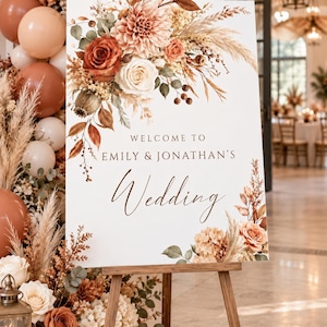 May include: A rectangular white wedding welcome sign on a wooden easel, featuring printed text that reads Welcome to Emily & Jonathan's Wedding, decorated with a floral arrangement of rust, cream, and beige flowers, pampas grass, and green leaves.
