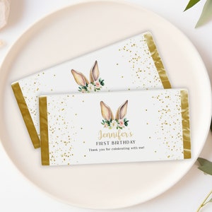 Può includere: Due involucri di barretta di cioccolato bianco con accenti di foglia d'oro. Gli involucri presentano un'illustrazione ad acquerello di orecchie di coniglio con fiori e glitter dorato. Il testo sull'involucro recita "Jennifer's First Birthday. Thank you for celebrating with me!"
