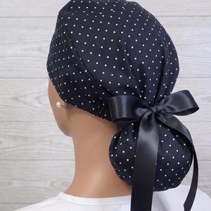 May include: A navy blue scrub cap featuring a white polka dot pattern. The cap has a gathered back design to accommodate hair and is secured with a smooth, solid black satin ribbon tied into a bow at the center of the back of the head.