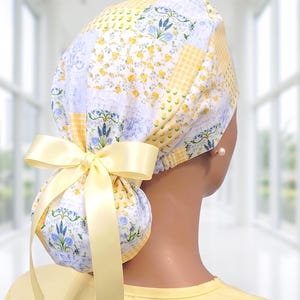 May include: A scrub cap featuring a patchwork design of yellow gingham, small yellow flowers, and blue lavender sprigs on a white background. The cap includes a gathered back section secured with a pale yellow satin ribbon tied into a bow.
