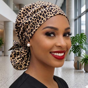 May include: A scrub cap featuring a tan fabric base with a black and brown leopard print pattern. The cap has a gathered design at the back with a tied knot detail, providing a secure fit around the head while covering the hair completely.
