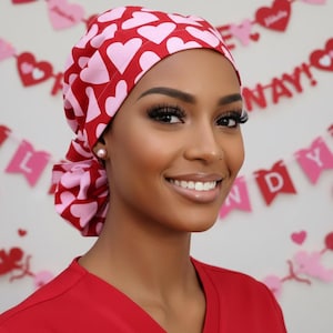May include: A red and pink heart-patterned scrub cap. The cap is made of fabric and features a repeating pattern of pink hearts on a red background. The cap is worn by a person wearing a red scrub top.