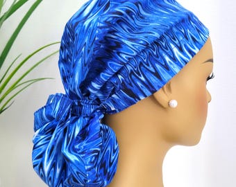 May include: A blue and white patterned scrub cap with a gathered ponytail holder. The cap is made of fabric with a swirling design in shades of blue and white. The cap is designed to be worn on the head.