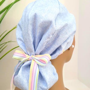 May include: A light blue denim scrub cap with a colorful striped bow. The cap is gathered at the back and tied with a matching ribbon. The bow features vertical stripes in pastel colors.