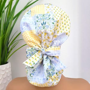 Pode incluir: Uma touca cirúrgica de patchwork com laço, feita de tecidos florais e xadrez em amarelo, azul e branco. O laço é amarrado na parte de trás. A touca é projetada para profissionais médicos, com vários padrões florais e xadrez.