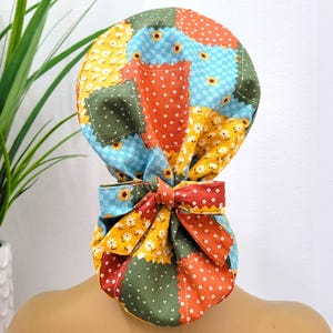 Op de afbeelding: Een kleurrijke patchwork scrub cap met een strik. De cap heeft een verscheidenheid aan stoffen in tinten blauw, geel, oranje en groen, met bloemen- en stippatronen. De strik is gemaakt van een bijpassende stof.