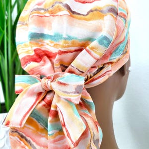 May include: A patterned headscarf with a bow. The fabric displays a watercolour design in orange, red, yellow, and teal. The bow is tied on the side, adding a decorative element to the headwear.