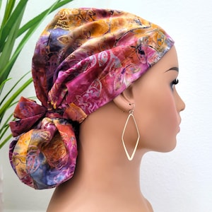 Puede incluir: Un pañuelo para la cabeza colorido con un patrón de estilo batik en tonos de rosa, morado, naranja y azul. El pañuelo está atado en la parte posterior. La modelo lleva pendientes dorados en forma de diamante.