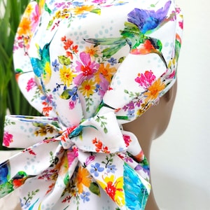 May include: A white scrub cap featuring a vibrant, multicolored pattern of hummingbirds and various wildflowers in shades of pink, orange, yellow, and blue. The cap includes a fabric tie at the back, allowing for an adjustable fit around the head.