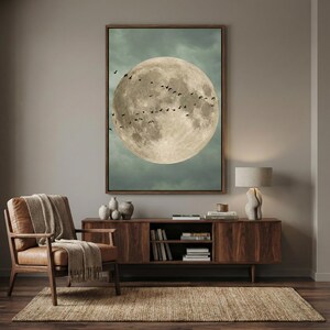 May include: A framed artwork depicts a large, detailed moon with a flock of birds in silhouette against a cloudy, teal sky. The artwork hangs above a wooden cabinet with decorative vases and a lamp.