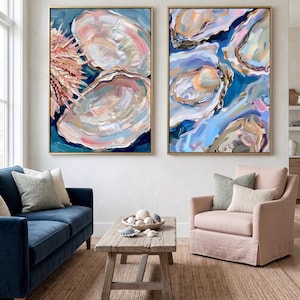 May include: Two framed abstract paintings with gold frames. The paintings depict oyster shells in shades of blue, pink, and cream. The artwork is displayed in a living room setting with a blue sofa, pink armchair, and wooden coffee table.