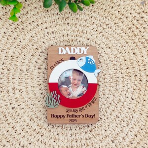 Peut inclure: Cadre photo en bois pour la fête des pères avec un design circulaire représentant un poisson bleu et blanc et le texte "DADDY". Le cadre comprend une photo de deux enfants et le texte "Happy Father's Day! 2025".