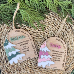 Puede incluir: Dos adornos navideños de madera con un diseño de árbol y el texto "These Are A Few Of My Favorite Things." Uno tiene un árbol verde y el nombre "Nolan", el otro un árbol rosa y el nombre "Tinky".