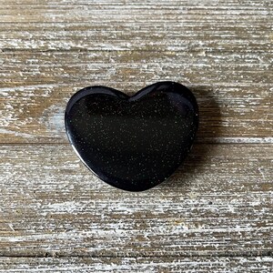 May include: A black heart-shaped decorative object with a glittery, speckled appearance. The heart is smooth and polished, reflecting light. The object is set against a weathered, light-colored wooden surface.