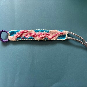 May include: A friendship bracelet with a blue, white, and pink woven pattern. The word "Forever" is woven in pink.
