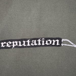 May include: Black and white friendship bracelet with the word "reputation" woven into the design. The bracelet has white tassels on the ends.