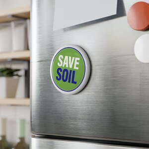 Könnte beinhalten: Ein runder Magnet in Grün und Weiß mit blauem Rand und dem Text "SAVE SOIL" in weißen Buchstaben.