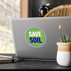 以下が含まれることがあります： 銀色のラップトップに「SAVE SOIL」という文字が書かれた緑と青の円形のステッカー