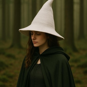May include: A woman wearing a cream-coloured witch hat with a wide brim and a pointed crown. She is also wearing a dark green hooded cloak and dress. The background is a blurred forest scene with tall trees.